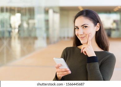 Cheerful Pensive Woman With Cellphone Touching Face With Finger And Smiling At Camera. Young Woman With Mobile Phone Posing Indoors With Glass Wall Interior In Background. Wireless Connection Concept