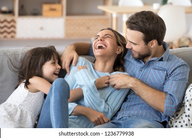 Cheerful people sitting on couch in living room have fun little daughter tickling mother laughing together with parents enjoy free time playing at home. Weekend activity happy family lifestyle concept