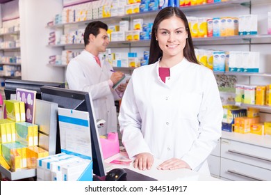 Cheerful Pharmacist Standing At Pay Desk And Pharmacy Technician Helping 