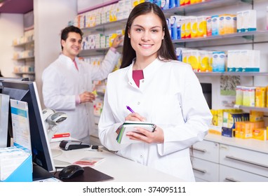 Cheerful Pharmacist Standing At The Pay Desk And Pharmacy Technician Helping 