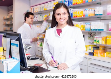 Cheerful Pharmacist Standing At Pay Desk And Pharmacy Technician Helping 