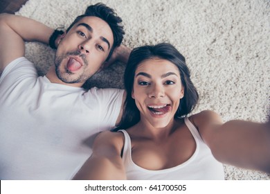 Cheerful Playful Hispanic Sister And Brother Are Taking Selfie And Making Funny Grimaces. They Lie On The Floor On Beige Carpet In White Casual Outfits Indoors At Home