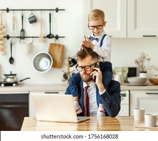 Cheerful playful kid sitting on neck of unhappy busy dad in formal wear during phone conversation and working with laptop in home kitchen