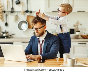 Cheerful Playful Kid Sitting On Neck Of Unhappy Busy Dad In Formal Wear During Phone Conversation And Working With Laptop In Home Kitchen
