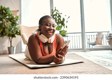 Cheerful plump woman with wireless headset on neck lying on yoga mat and using smartphone. Black lady in sport clothes having break during home sport with gadget in hands. - Powered by Shutterstock