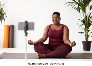 Cheerful plus size young black woman in nice sportswear doing yoga at home, sitting on fitness mat in lotus position and looking at smartphone screen, smiling, having online yoga class - Powered by Shutterstock