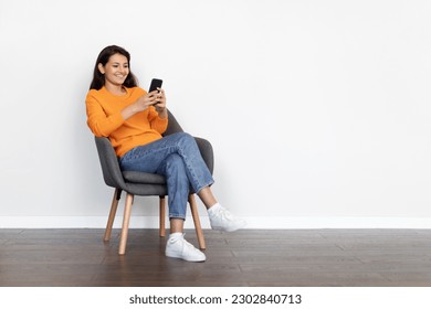 Cheerful positive attractive young indian woman wearing casual outfit sitting in armchair at home, using cell phone, chatting with guys on dating app, shopping online, scrolling, copy space