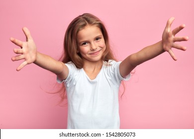 cheerful positive beautiful girl in white t-shirt with arms wide open looking at the camera, kid meeting her guests, welcome, kindness, friendship. close up portrait, isolated pink background