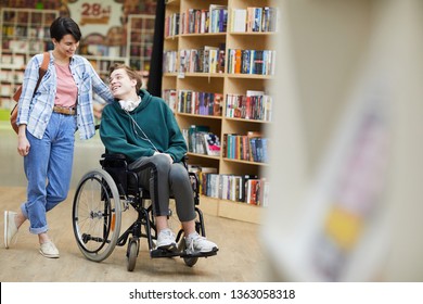 Cheerful positive handsome disabled student with headphones on neck sitting on wheelchair and talking to groupmate while they visiting library together