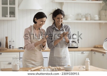 Cheerful positive mature mom and adult child woman baking pies, biscuits, shaping balls from raw dough at kitchen table with bakery ingredients, laughing, enjoying household cooking activities