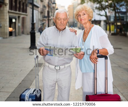 Cheerful positive  senior couple of tourists with map and city guide walking on street 