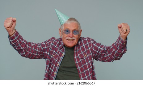 Cheerful Positive Surprised Senior Man In Cone Festive Hat Cap, Dancing, Having Fun, Celebrating Birthday Party Event Holiday, Anniversary. Mature Old Grandfather Alone On Gray Studio Wall Background