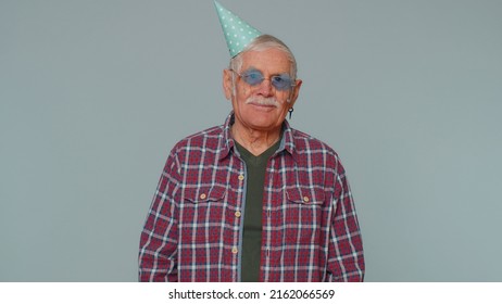 Cheerful Positive Surprised Senior Man In Cone Festive Hat Cap, Dancing, Having Fun, Celebrating Birthday Party Event Holiday, Anniversary. Elderly Old Grandfather Alone On Gray Studio Wall Background