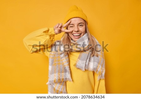 Cheerful positive woman makes peace gestue over eye winks and smiles gladfully gestures victory sign wears casual jumper scarf around neck and hat isolated over yellow background. Body language