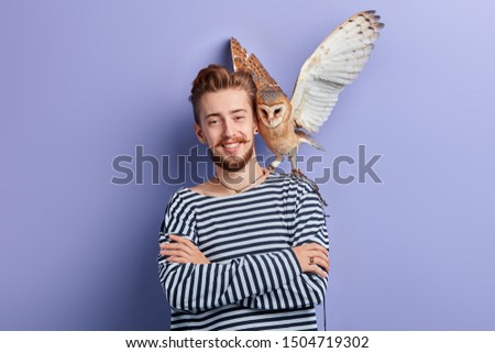 cheerful positive young man with an owl in his shoulder which is going to fly. isolated blue background, studio shot.