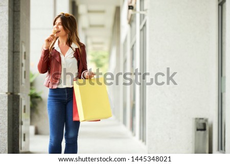Cheerful positive young woman in casual clothes enjoying tasty icecream when walking outdoors with many shopping bags