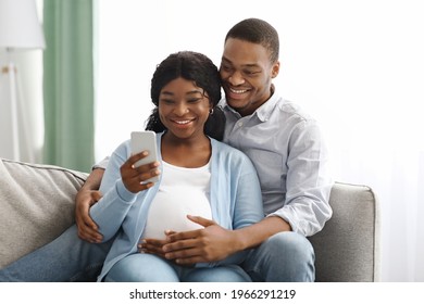 Cheerful pregnant african american couple using mobile phone while resting together on couch in living room, copy space. Happy expecting black family checking mobile applications for future parents