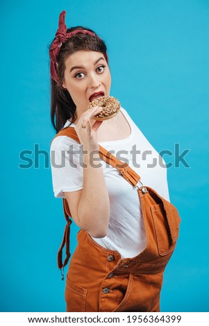 Cheerful pregnant brunette woman eating donuts dressed in pin-up style isolated on blue background. Happiness from pregnancy while expecting a baby. High quality photo