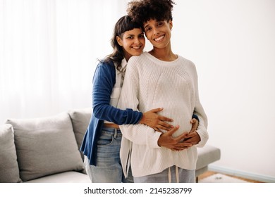 Cheerful pregnant couple smiling at the camera while standing indoors. Happy young woman embracing her pregnant wife from behind. Pregnant lesbian couple feeling excited about their unborn baby.