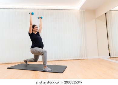 A cheerful pregnant doing lunges with light dumbells exercises on a mat at health center - Powered by Shutterstock