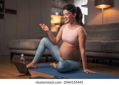 A cheerful pregnant lady explaining a home workout exercise on her live stream while using a tablet and sitting on an exercise mat in her living room. - Powered by Shutterstock