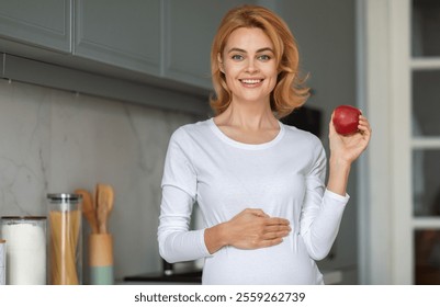 A cheerful pregnant woman stands in a bright kitchen, holding a red apple while gently cradling her baby bump. The warm atmosphere highlights her joy and anticipation. - Powered by Shutterstock