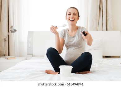 Cheerful pregnant young woman with ice cream laughing and watching TV on bed at home