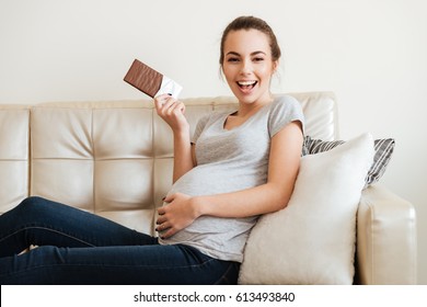 Cheerful Pregnant Young Woman Sitting On Sofa And Eating Chocolate At Home