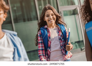 Cheerful preteen caucasian girl elementary middle school pupil with backpack and book in casual clothes going from school and looking at camera together with her friends classmates - Powered by Shutterstock