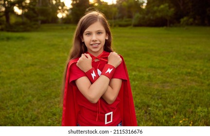 Cheerful Preteen Child Wearing Bright Red Hero Costume Showing Superpower While Standing With Crossed Hands On Chest And Looking At Camera In Summer Park