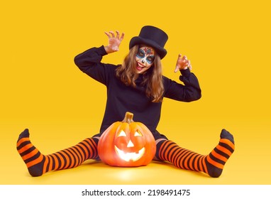 Cheerful Preteen Girl With Halloween Makeup Makes Scratching Gesture On Orange Background. Child With Creative Makeup Sits Near Halloween Pumpkin And Scares You By Waving His Arms On Orange Background