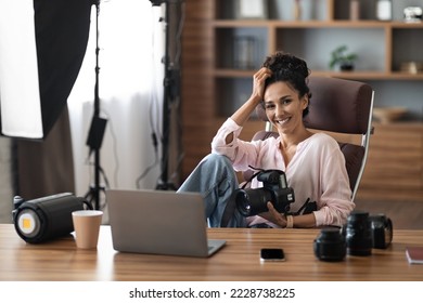 Cheerful pretty brunette young woman in casual outfit professional photographer posing at workplace photo studio, sitting at desk in front of modern laptop, holding camera, copy space - Powered by Shutterstock