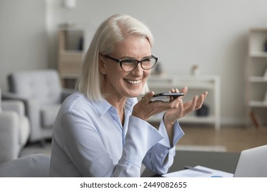 Cheerful pretty businesswoman sit at workplace, speak on speakerphone on mobile phone, record audio message, chatting, share messages, enjoy pleasant conversation, use virtual assistant application - Powered by Shutterstock