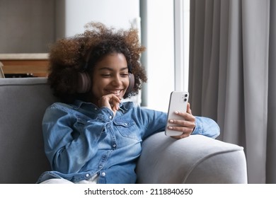 Cheerful pretty gen Z African teen girl listening to popular music, using smartphone, big headphones, looking at mobile phone screen, smiling, talking on video call, watching media
