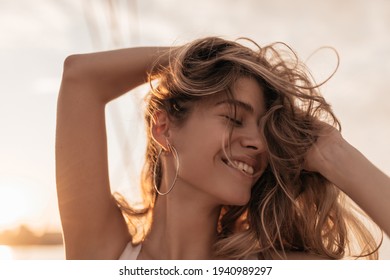 Cheerful pretty girl with closed eyes, long blonde hair, stylish accessories smiling and posing against background of pink sunset sky - Powered by Shutterstock