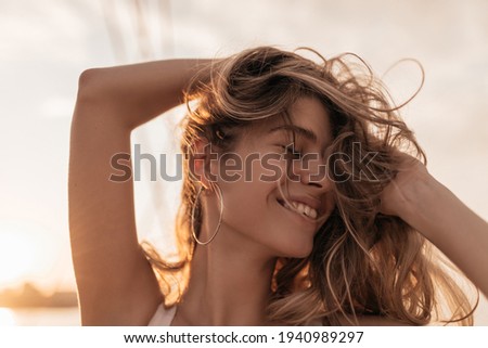 Cheerful pretty girl with closed eyes, long blonde hair, stylish accessories smiling and posing against background of pink sunset sky