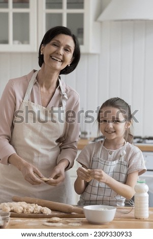 Cheerful pretty grandmother and sweet kid girl in aprons baking dessert t kitchen table together, shaping dumplings, pies, cookies over floury table, looking at camera, smiling, laughing