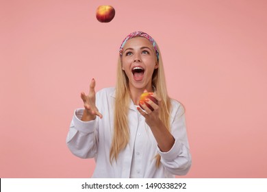 Cheerful pretty lady with long blond hair juggling with peaches in studio, having fun over pink background, wearing colored headband and white shirt