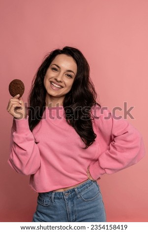 Cheerful pretty lady with an oat biscuit in one hand posing for the camera on the pink background. Self-indulgence and unhealthy food concept