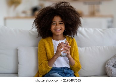 Cheerful Pretty Little Black Girl With Bushy Hair Sitting On Couch At Home, Holding Glass Of Water And Smiling At Camera, Kid Drinking Water, Copy Space. Hydration, Children Healthy Lifestyle