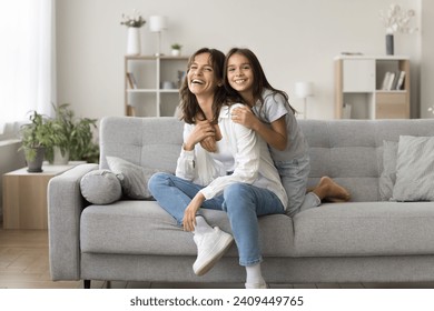 Cheerful pretty loving little girl hugging happy young mom on home sofa, spending leisure with mother at comfortable home, enjoying family time in modern cozy interior, looking at camera for portrait - Powered by Shutterstock