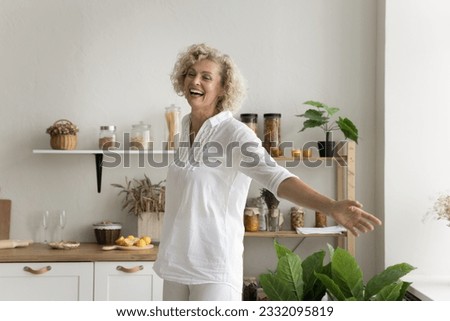Cheerful pretty mature woman having fun at home kitchen, dancing with open arms, laughing, enjoying music, active leisure time, feeling happy, carefree, inspired