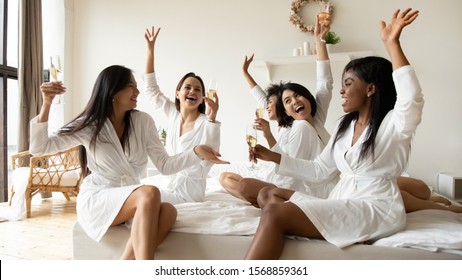 Cheerful pretty multiracial young ladies friends wear silk robes having fun hold glasses with champagne sit on bed in hotel, happy diverse girls bride and bridesmaid celebrate bachelorette spa party