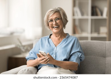 Cheerful pretty older retired woman in stylish glasses holding smartphone on couch, looking at camera, smiling with white healthy teeth, laughing, posing for home portrait with digital gadget - Powered by Shutterstock