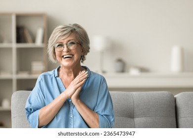 Cheerful pretty older woman in elegant glasses sitting on cozy home couch, smiling with perfect white teeth, laughing with hands at chest gesture, enjoying leisure, comfort, having fun
