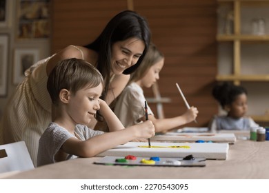 Cheerful pretty teacher girl helping pupil boy with drawing picture on canvas, standing at kid, smiling, laughing, looking at canvas. Child attending artistic school class, learning painting - Powered by Shutterstock