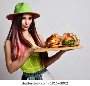 Cheerful pretty woman with pink long hair and bright make up wearing sexy green costume with hat standing and holding tray with fresh burgers over white background. Stylish casual look, fast food