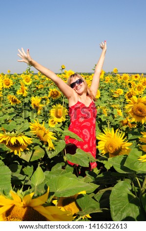 Cheerful pretty woman stands among blooming sunflowers with raised hands up to clear blue sky. Blonde hair woman in sunglasses and red dress with yellow flowers background. Summer vibes.