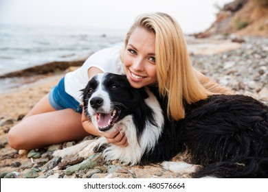 Cheerful Pretty Young Blonde Woman Sitting And Hugging Her Dog On The Beach