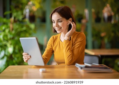Cheerful pretty young brunette woman SMM manager working at cafe, freelancer sit at table at green cozy lounge, using modern digital pad and wireless earpods, have video call with client, copy space